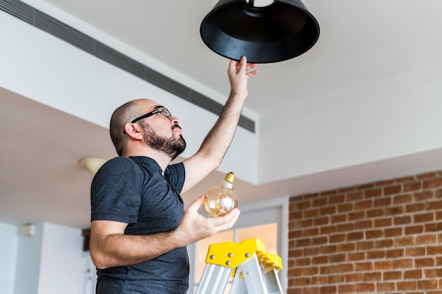Мужчина стоит на лестнице и заменяет лампочку в потолочном светильнике.